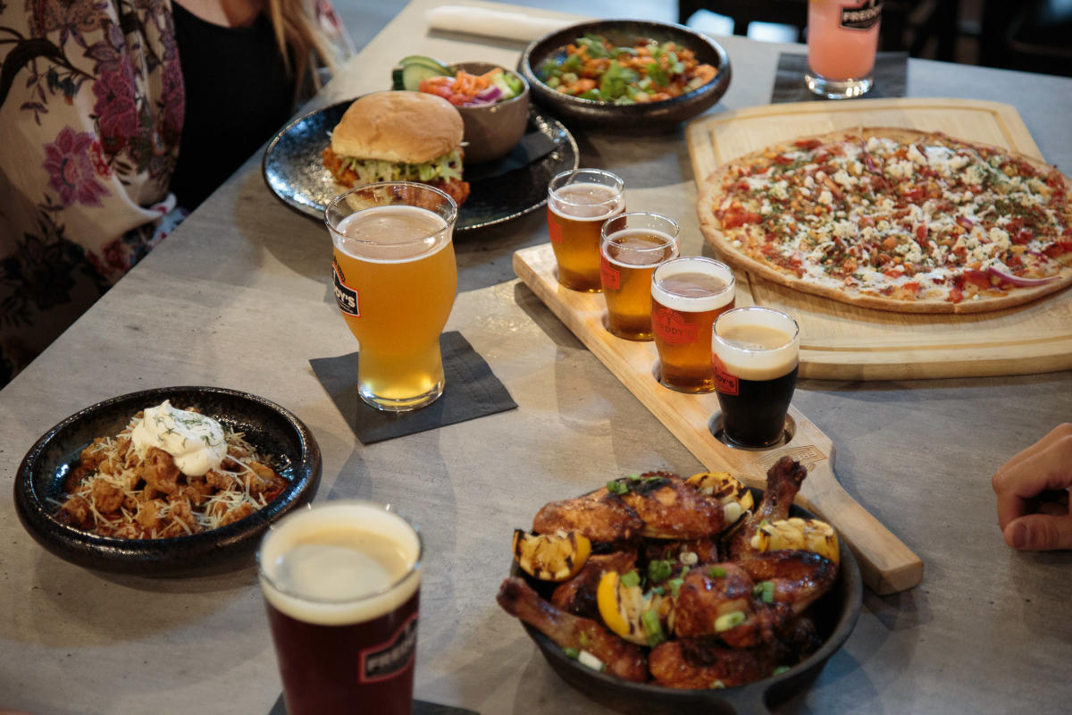 A table is filled with food and drinks, including a beer flight, a large pizza on a wooden cutting board, a sandwich, and a chicken salad at Freddy's BrewPub