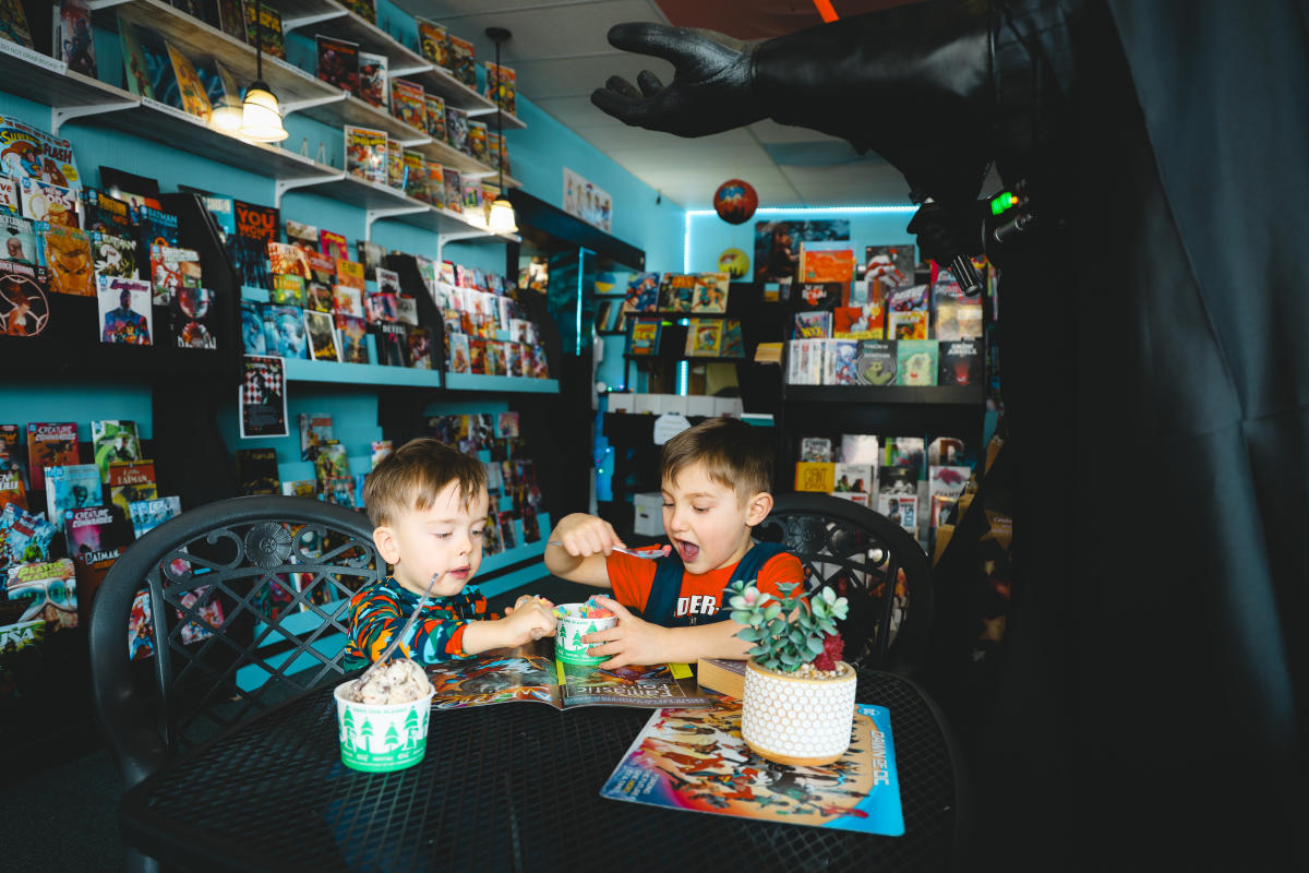 two kids enthusiastically eat ice cream from cups at a table inside a comic book store