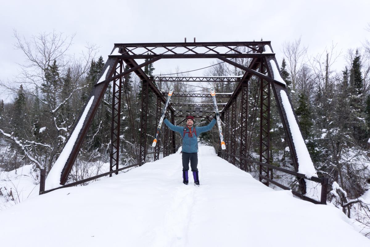 Man holds skis in air on old metal bridge.