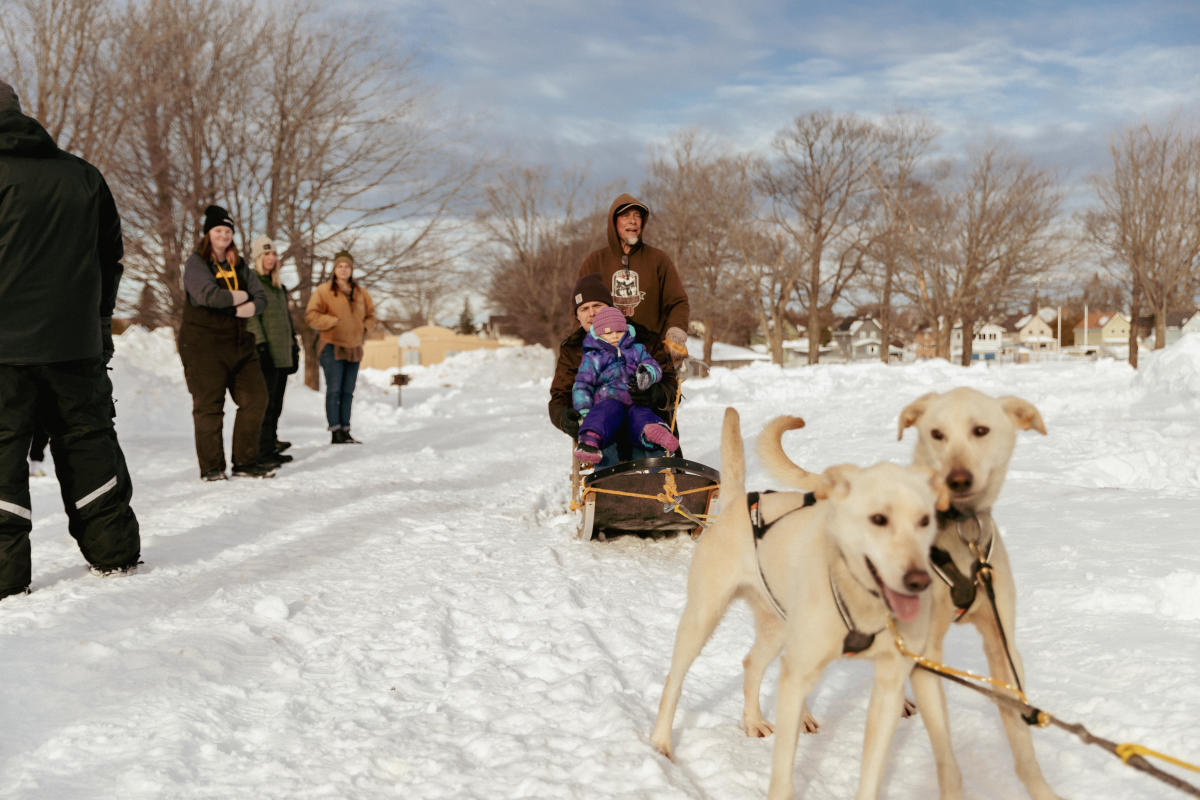 an adult holds a child as a musher drives a dog sled