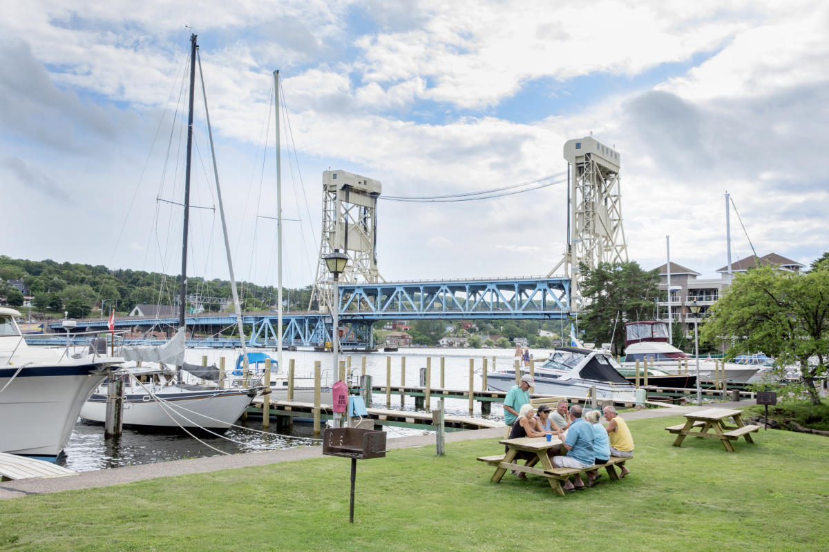 A group of people sit at a picnic table at the Houghton County Marina with the Portage Lake Lift Bridge in background.