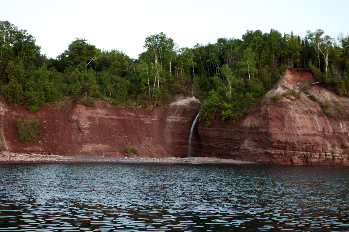 Waterfall spills over sandstone cliff onto Lake Superior beach.