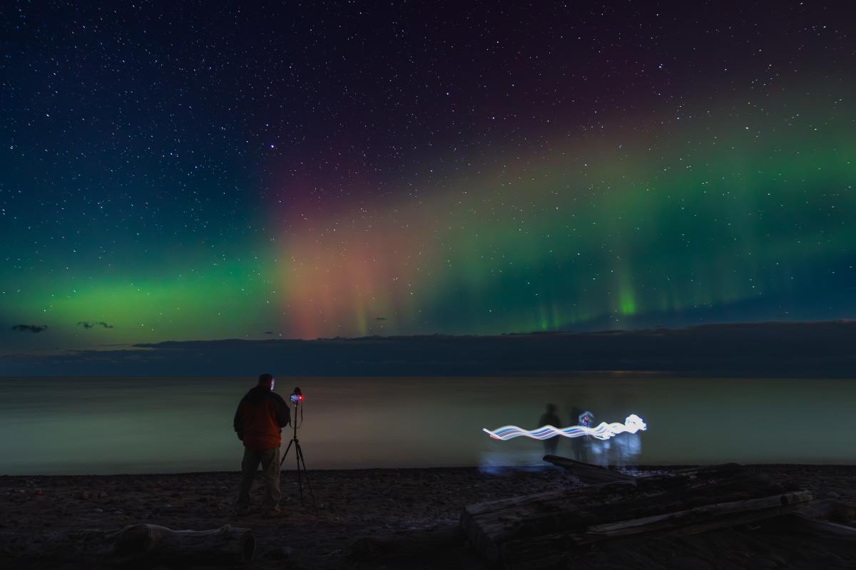 aurora dances over a lake while photographers await, some creating additional light pollution
