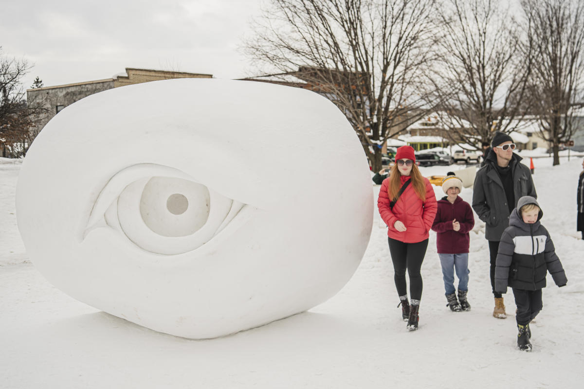 two parents and two children walk together in snow gear next to a snow statue of a human eye taller than an average person