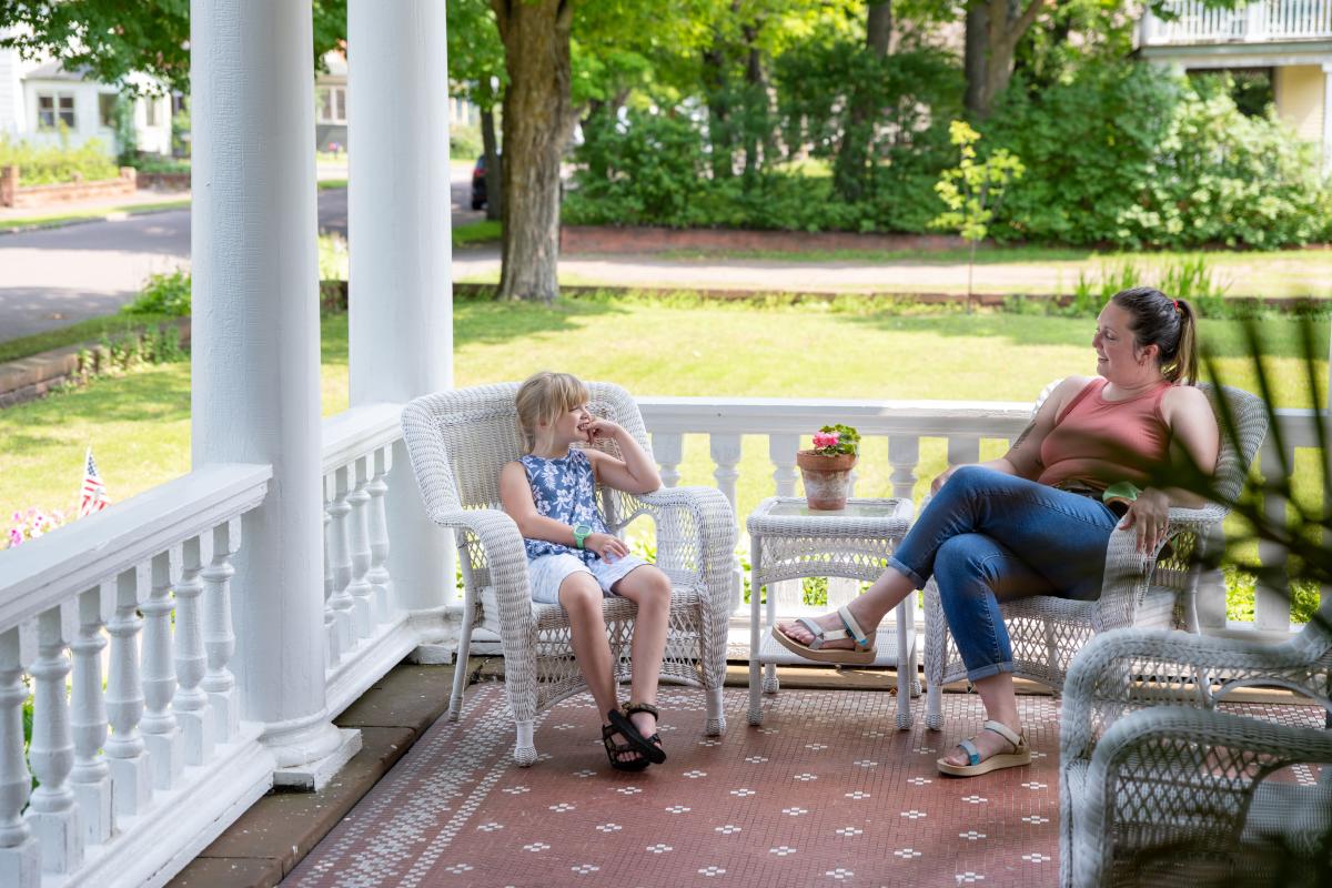 a child sits in a wicker chair across from an adult on a wide porch