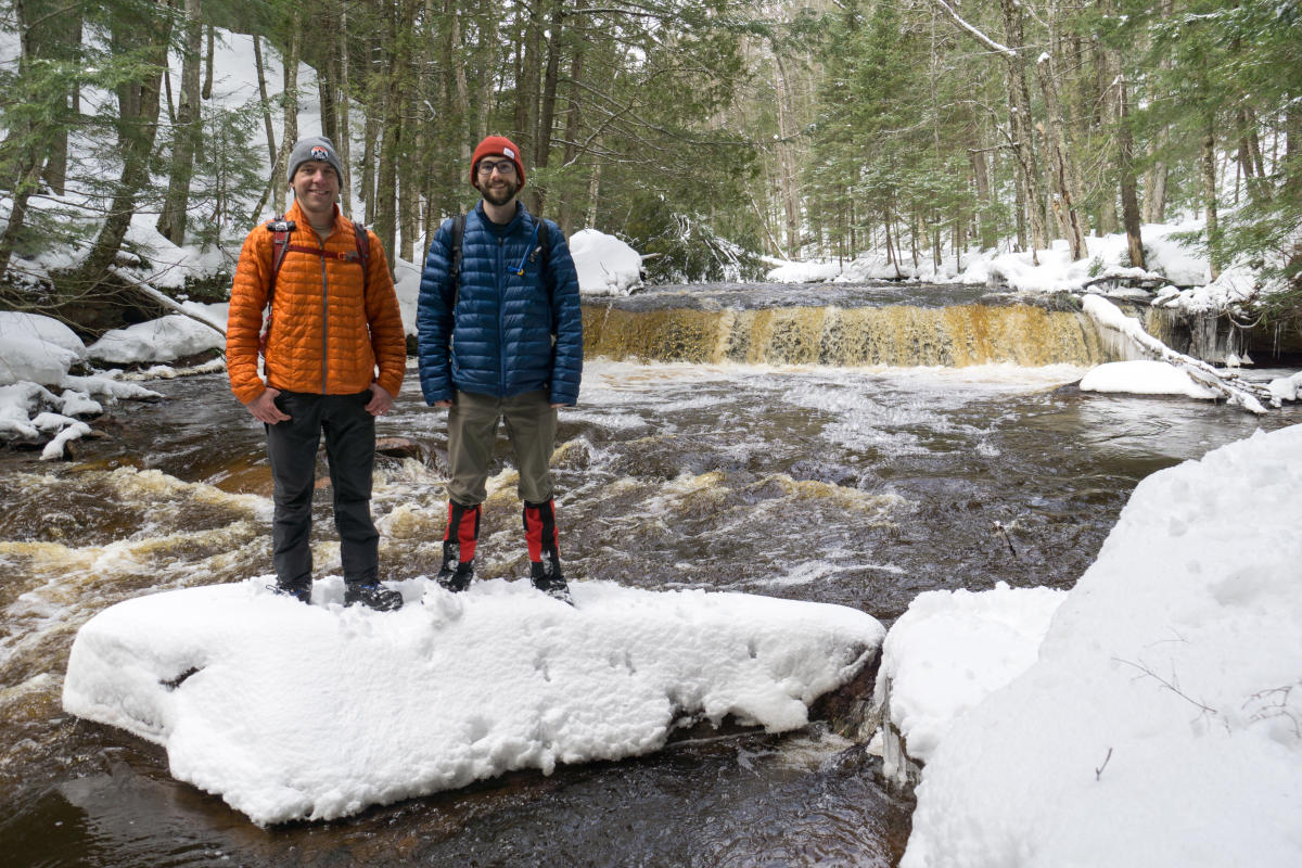 Two men smile for the camera on a snowy rock down stream from West Branch Falls. The falls can be seen in the background.