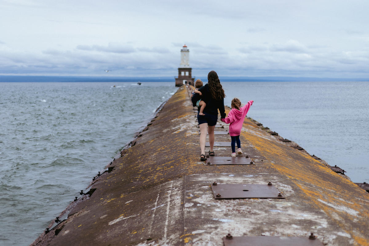 an adult carries a child and hold the hand of a toddle as they walk along a pier towards a lighthouse