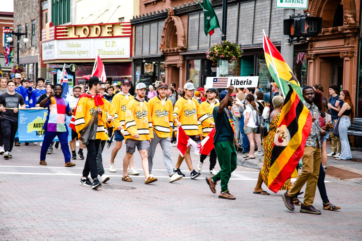 a parade of people waving international flags walks down a historic small town street