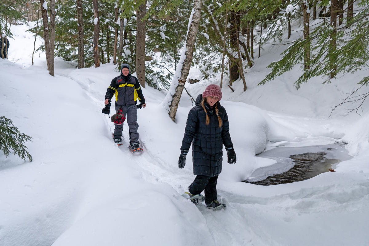 Two snowhoers walk along a path near the river.