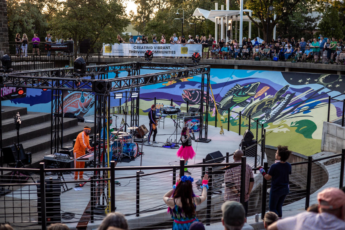 A band performs at Fish Ladder Music Park, with a crowd of people in stands and looking down toward the stage from the fence surrounding the fish ladder. There's a large colorful mural on the side of the structure showing fish with headphones and musical instruments.
