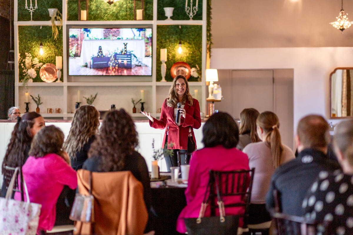 A woman speaks in front of a group of people sitting at tables.