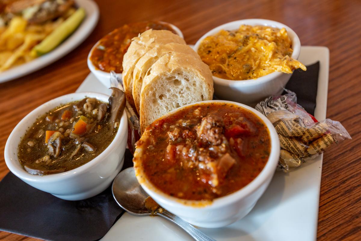 A plate holding four bowls of different soups, crackers, bread, and a spoon.