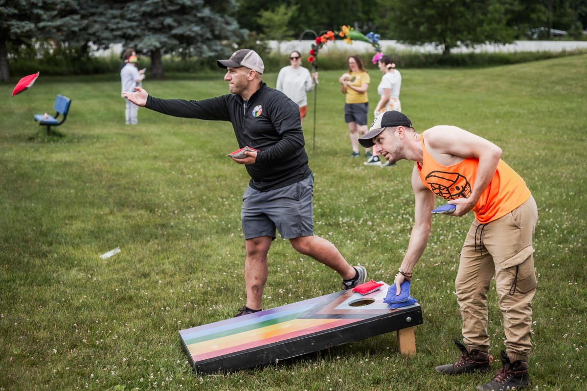 Two men playing cornhole at a Pride event in Mason. There are rainbow colors on the cornhole board and people in the background.
