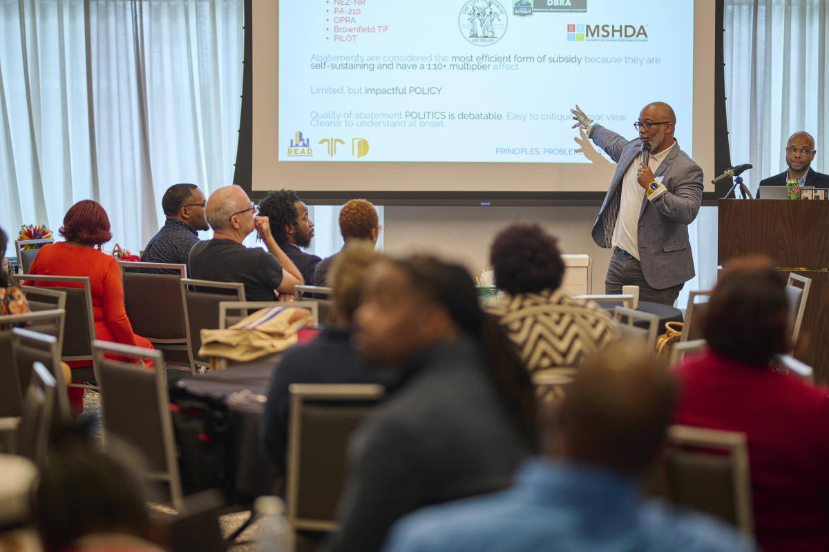 Numerous people facing towards a video screen in a conference room as the presenter is pointing to something on the screen