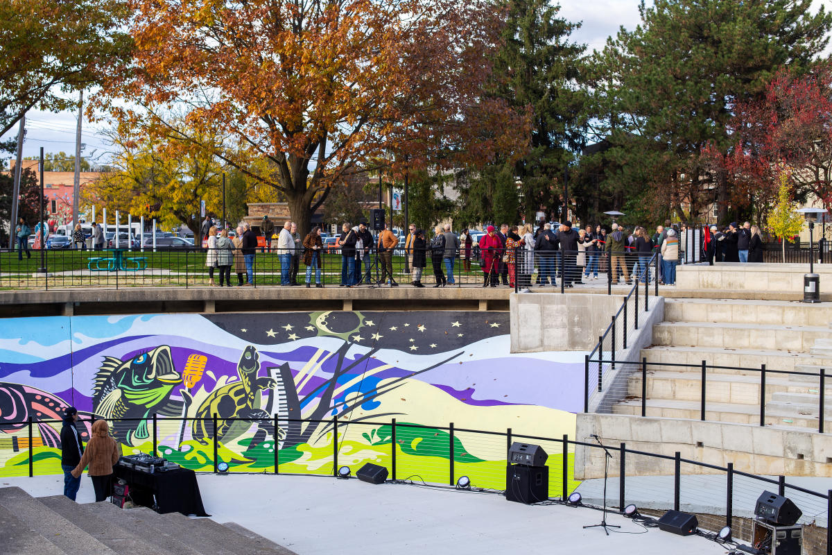 The stage, mural and some of the seating at the Fish Ladder Music Park with a crowd of people standing around