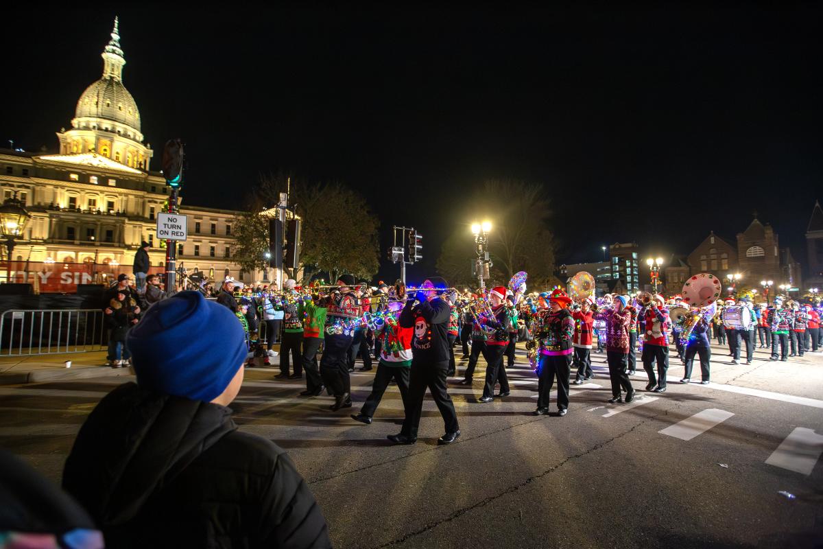 A marching band with lights parades in front of the Michigan State Capitol building while a crowd of people watch.