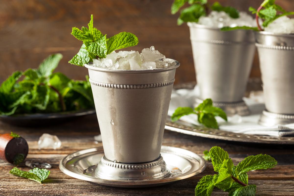 Mint Julep cocktail in an antique, silver cocktail glass sits on a silver platter with fresh sprigs of mint in the drink and on the wooden table.