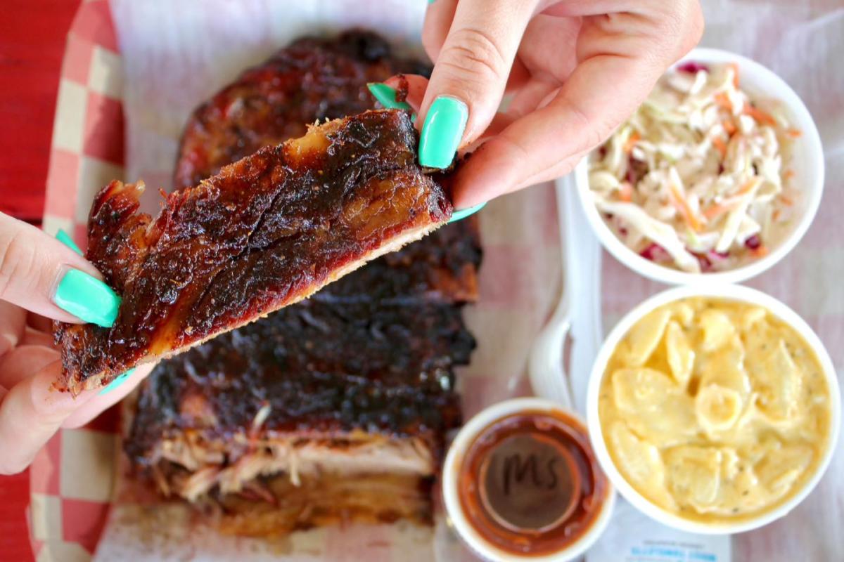 A hand holds a BBQ rib. Coleslaw and mac and cheese side dishes below.