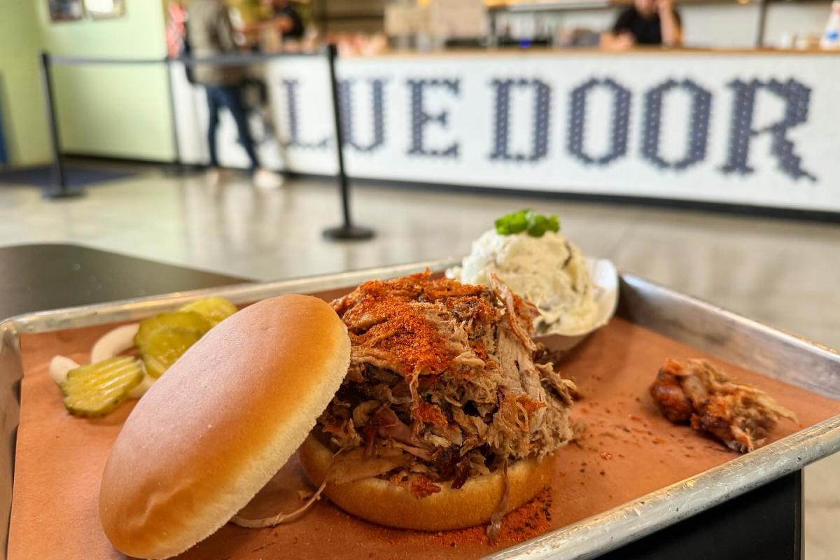 A pulled pork sandwich with potato salad and pickles in the background.