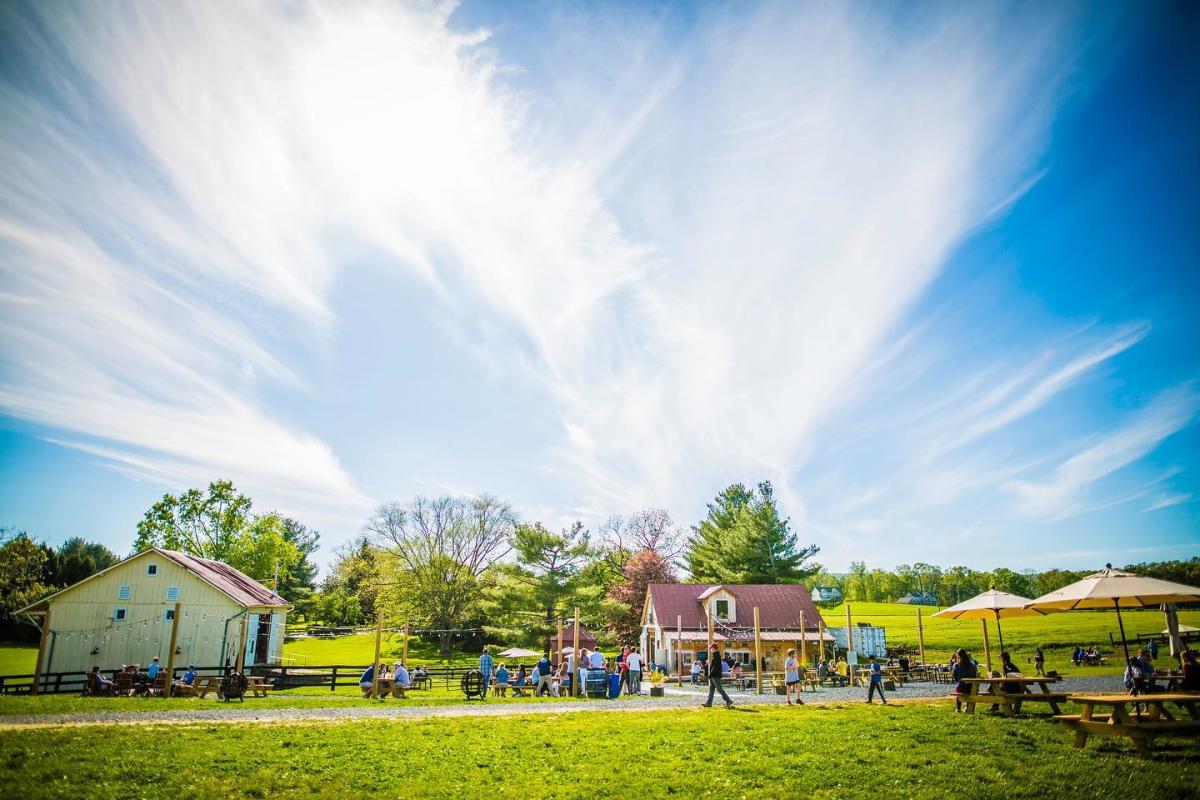 Families gather in the outdoor seating area at Wheatland Spring with a small barn on the left, a larger structure in the center of the photo and picnic tables lining a stone path that runs the length of the photo