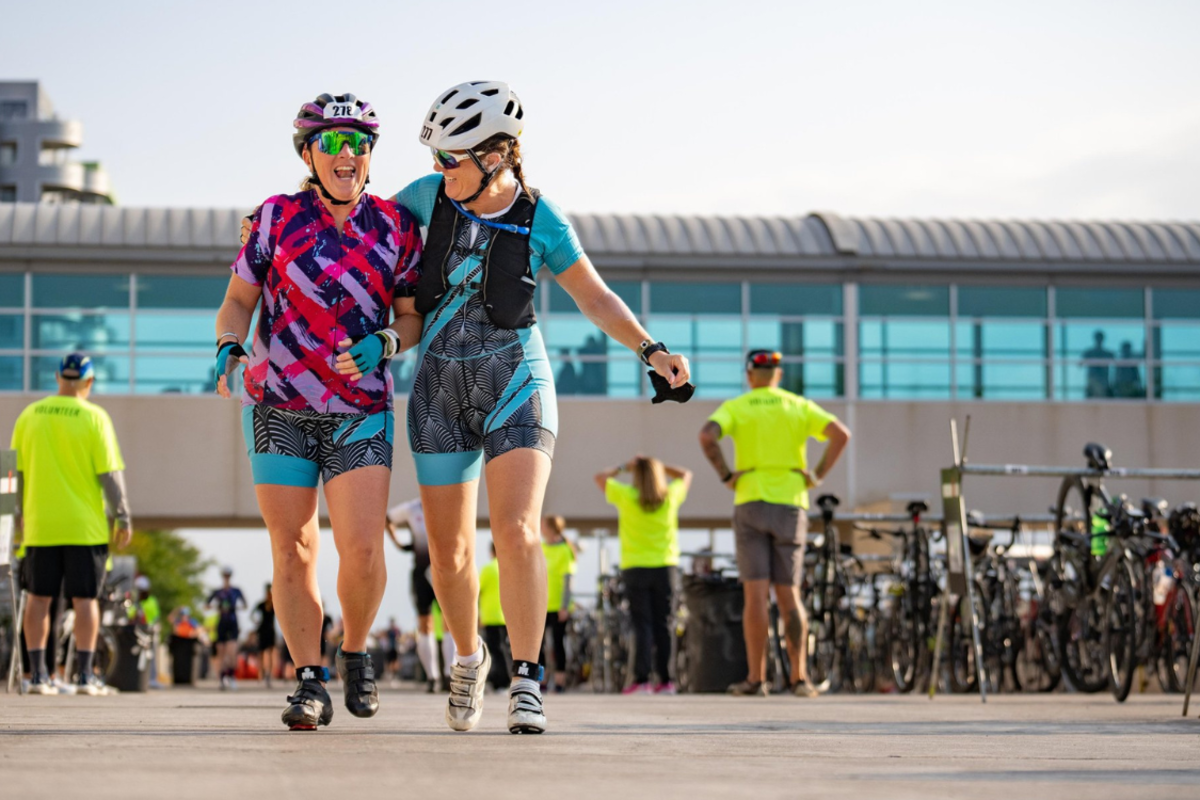 Two woman athletes wrap their arms around each other as they smile and walk away from IRONMAN Wisconsin bike transition area