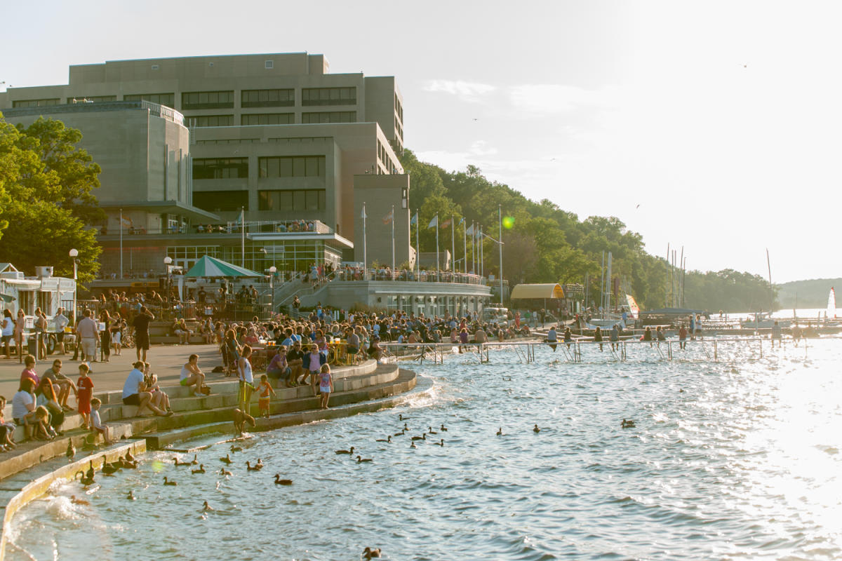 A view of Memorial Union Terrace from the Lake Mendota shoreline. The sun is setting on a summer day and dozens of people are along the shoreline looking at the lake which is dotted with dozens of ducks.