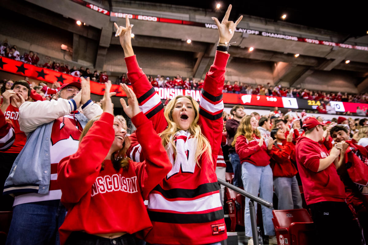 A white woman yells and raises her hands in the air in the crowd at a men's hockey game at the Kohl Center. She is wearing a red Badgers hockey jersey.