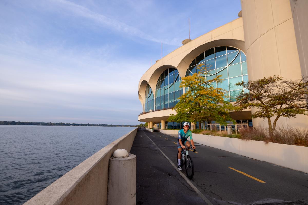 A man in a biker suit wearing a helmet as he rides his bike past the lake and Monona Terrace Convention Center.