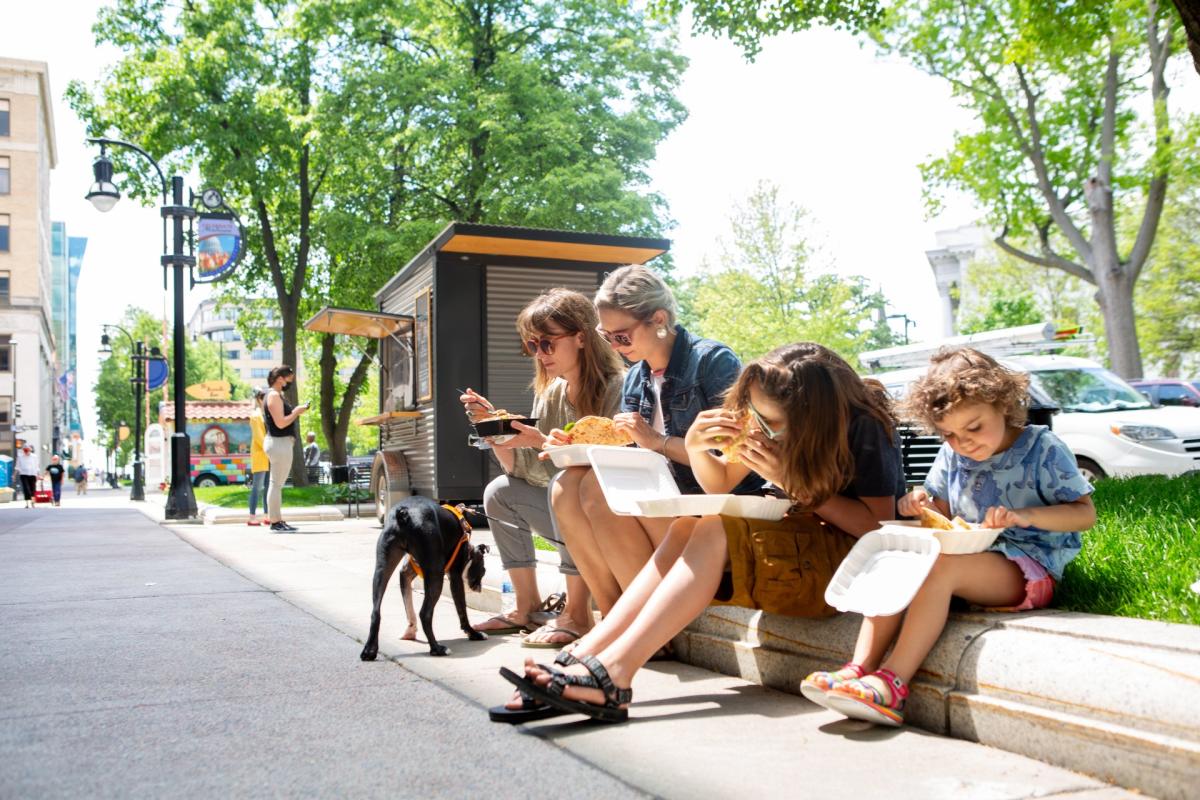 A family enjoys food from a food cart while sitting on a curb in downtown Madison