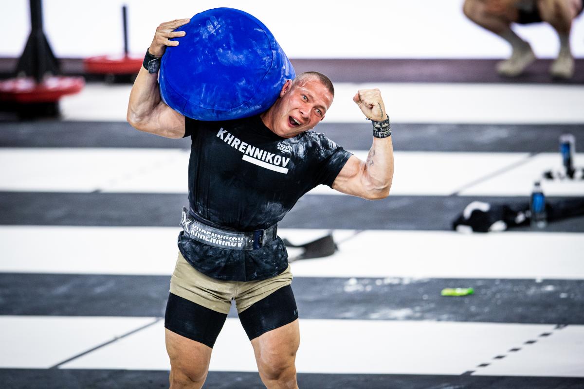 A muscular white man holds a bright blue sandbag on his right shoulder and flexes his left bicep at the CrossFit games. He's smiling triumphantly.
