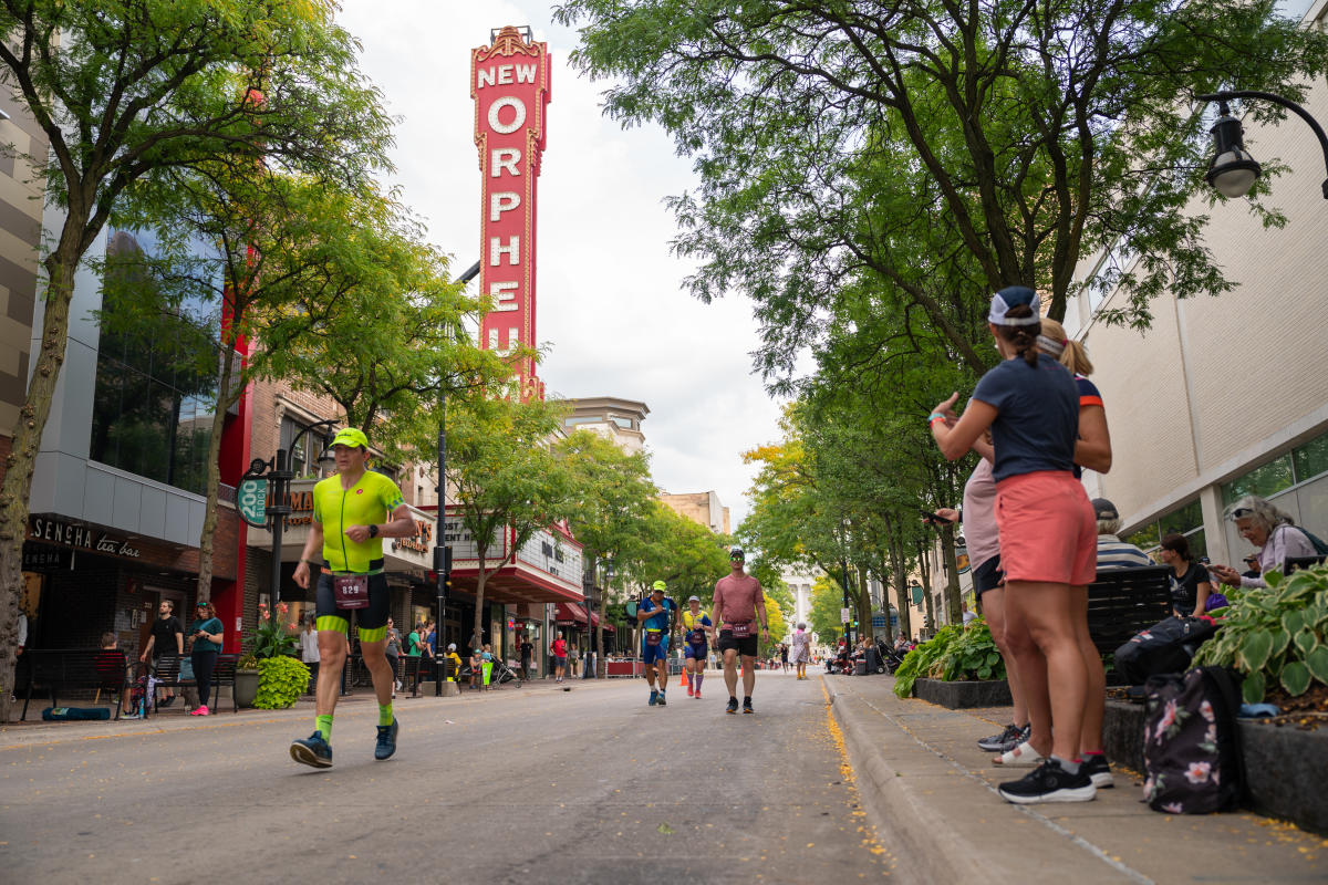 A group of five IRONMAN competitors run up State Street with the large, red marquee Orpheum sign in the background and green foliage surrounding them