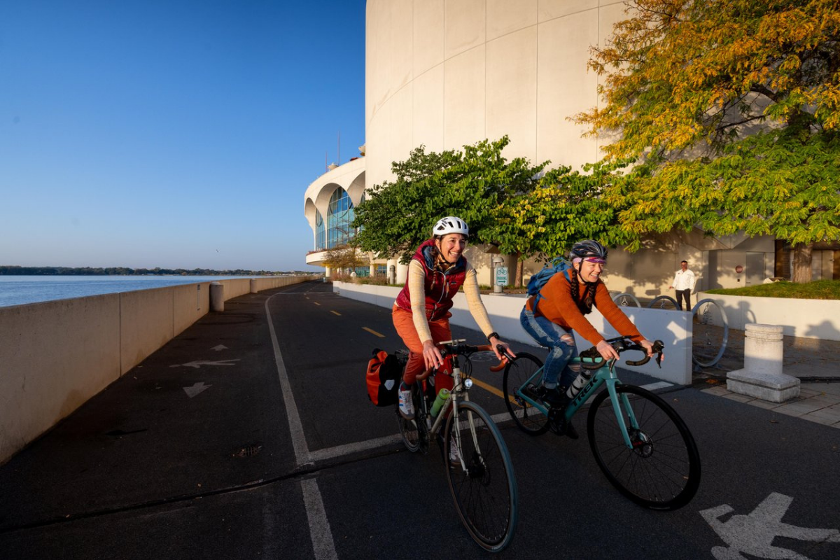 Two women on bikes biking Lake Monona in Madison with the Monona Terrace building in the background and fall foliage