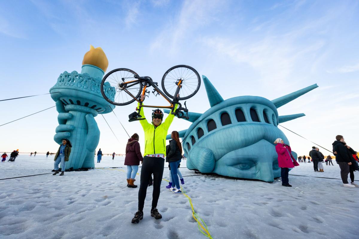 A man in biking clothes holds a bike over his head while standing on a frozen lake in front of a giant Lady Liberty inflatable statue