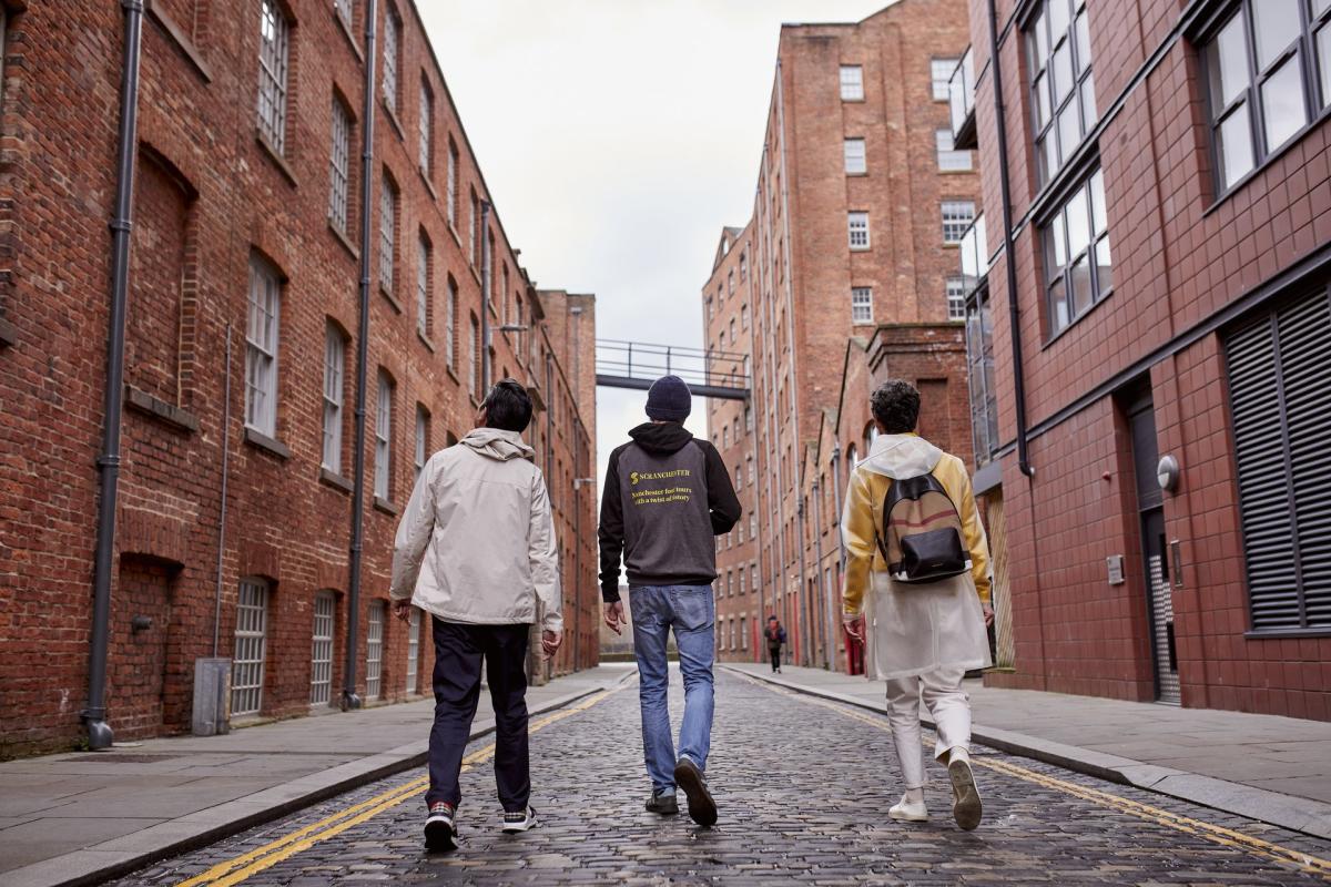 people walking on cobbled street with red brick mills in Manchster