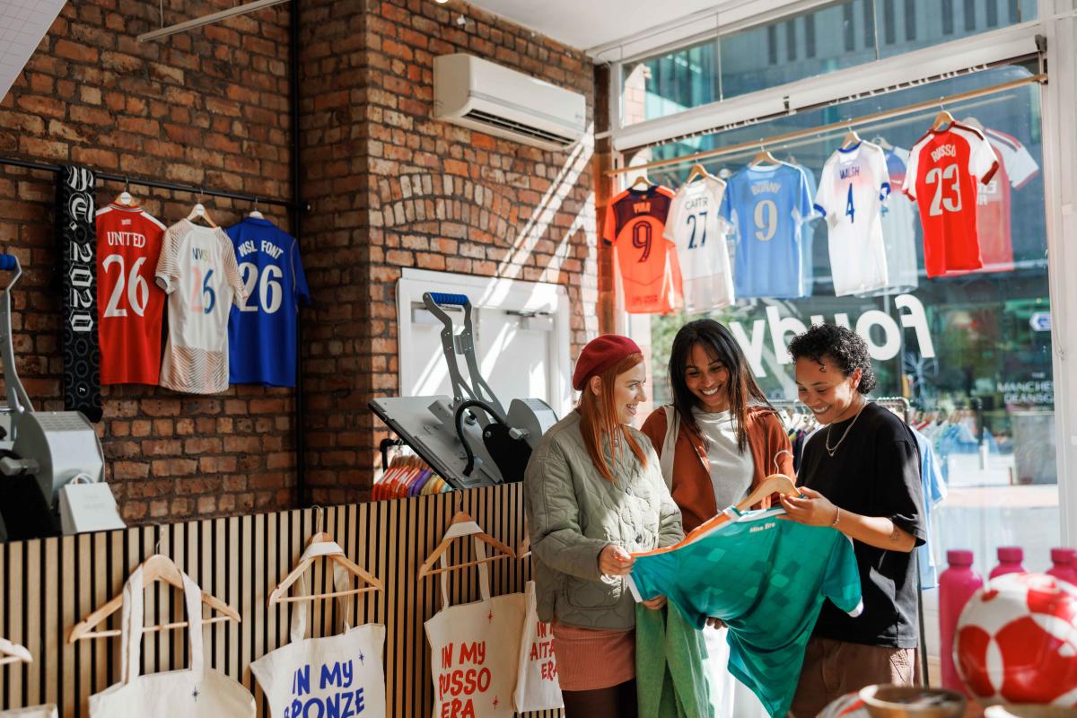 Three people browse football shirts inside a sportswear and memorabilia shop. The store features exposed brick walls, framed football jerseys, and clothing displays, while additional shirts are visible hanging in the window facing the street outside.