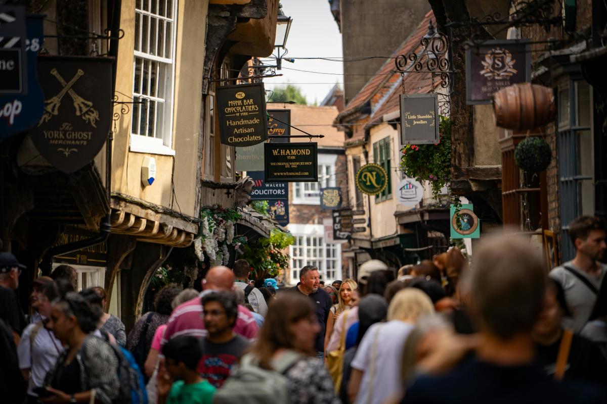 Crowded narrow street lined with historic timber-framed buildings and hanging shop signs, with many people walking beneath signs for shops including chocolatiers and jewellers, creating a busy market atmosphere.