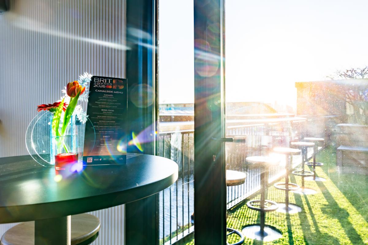 Interior table at Canalside, Co-op Live, with a small vase of flowers and a BRIT Awards 2026 Canal Side menu placed by a window overlooking the outdoor terrace and canal, sunlight streaming in.