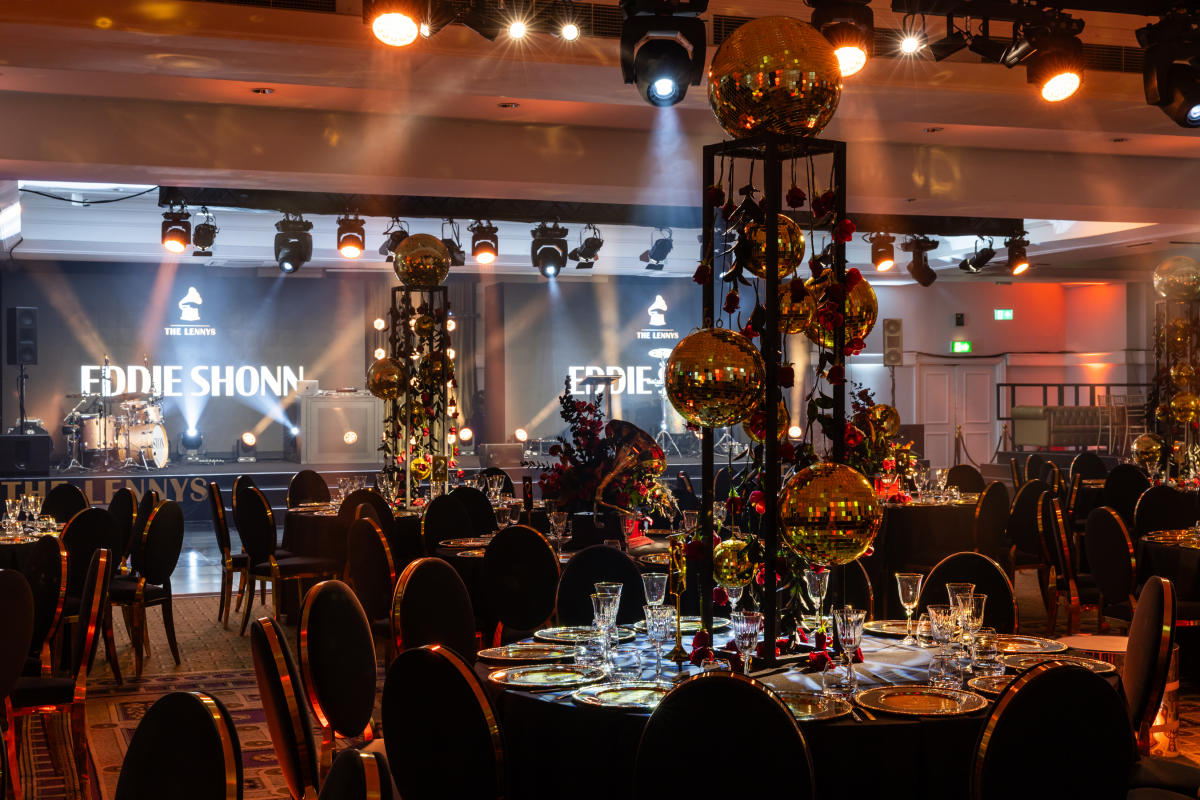Festive banquet hall set for a Christmas event, with round tables decorated with gold ornaments and centrepieces, stage lighting in the background, and rows of chairs arranged for guests.