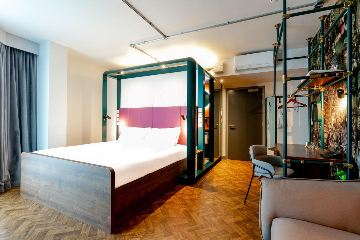 Interior of a YOTEL hotel bedroom, featuring a modern double bed with a framed headboard, wooden flooring, a small desk and seating area, and contemporary shelving and lighting.