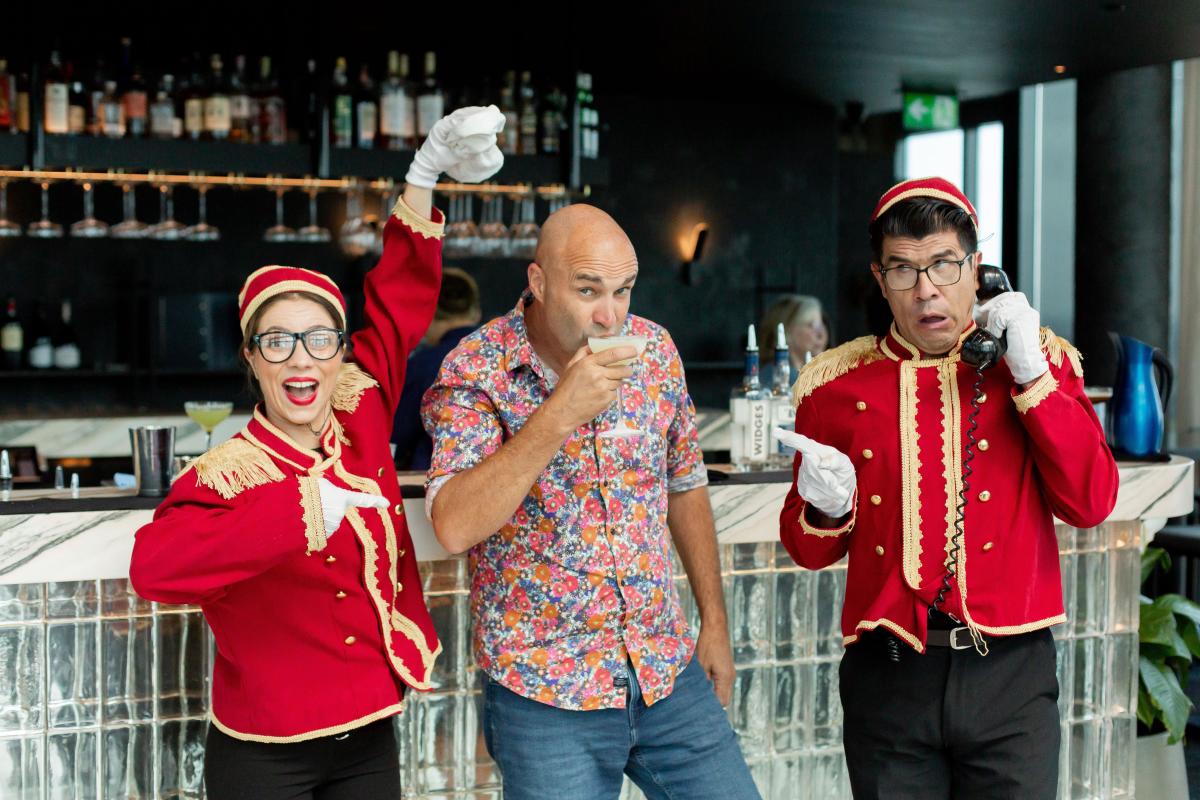 Two comedic entertainers in red theatre-usher style coats posing at the bar with a man drinking a glass of wine.