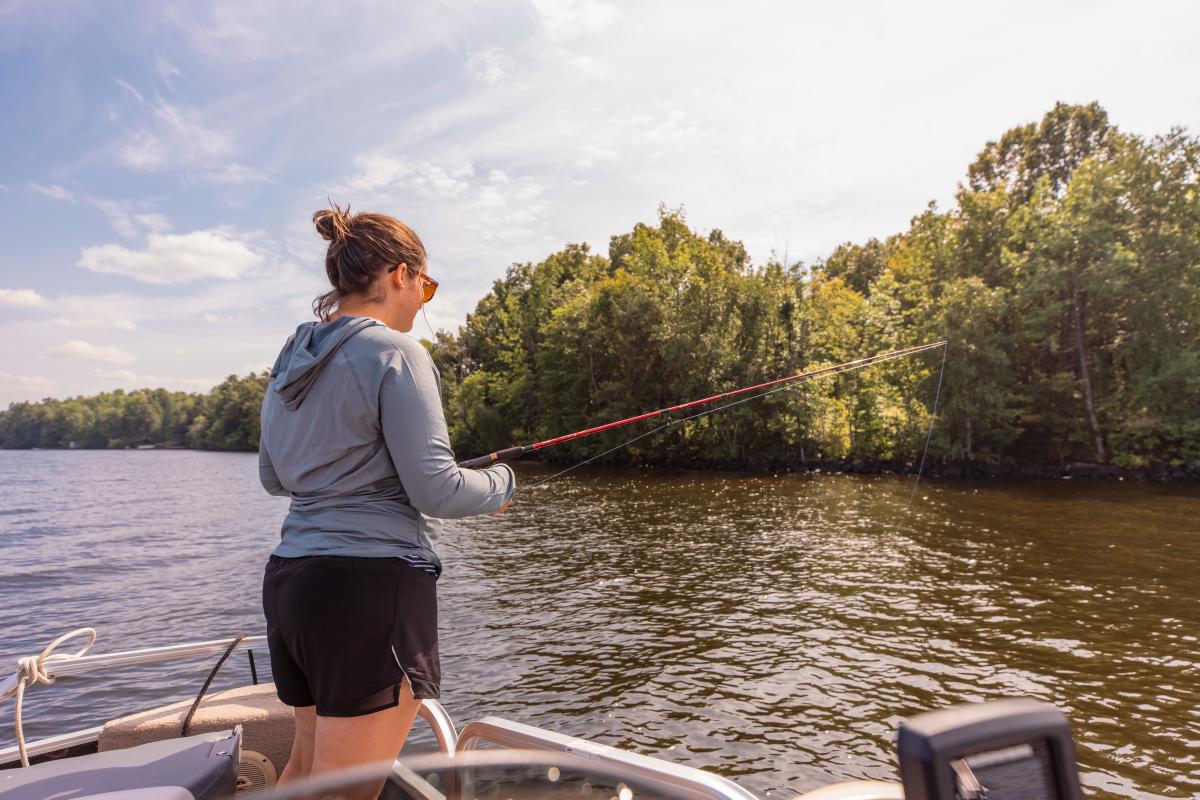 A woman fishes from a boat on a calm Lake Gogebic under a partly cloudy sky. She wears sunglasses and a hoodie, surrounded by lush green trees.