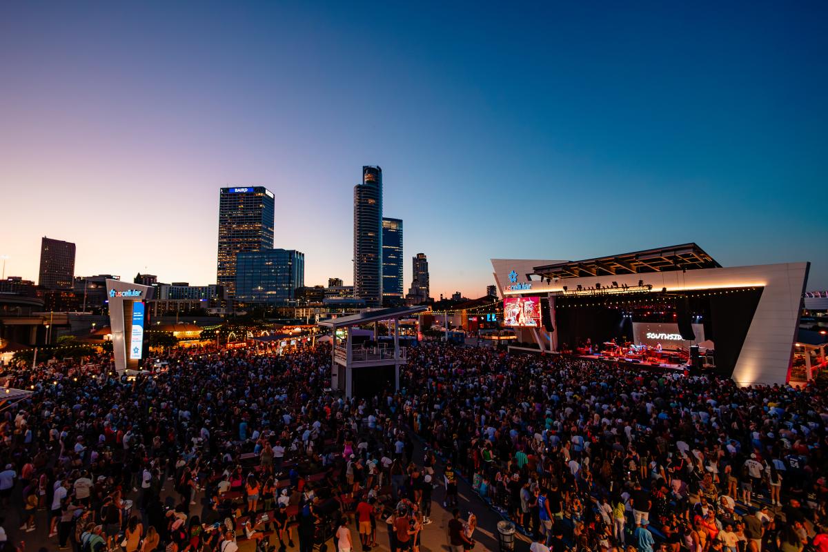 A large crowd gathers at the US Cellular Connection Stage during Summerfest in Milwaukee as the sun sets behind the city skyline, with high-rise buildings including the Baird Center visible in the background.