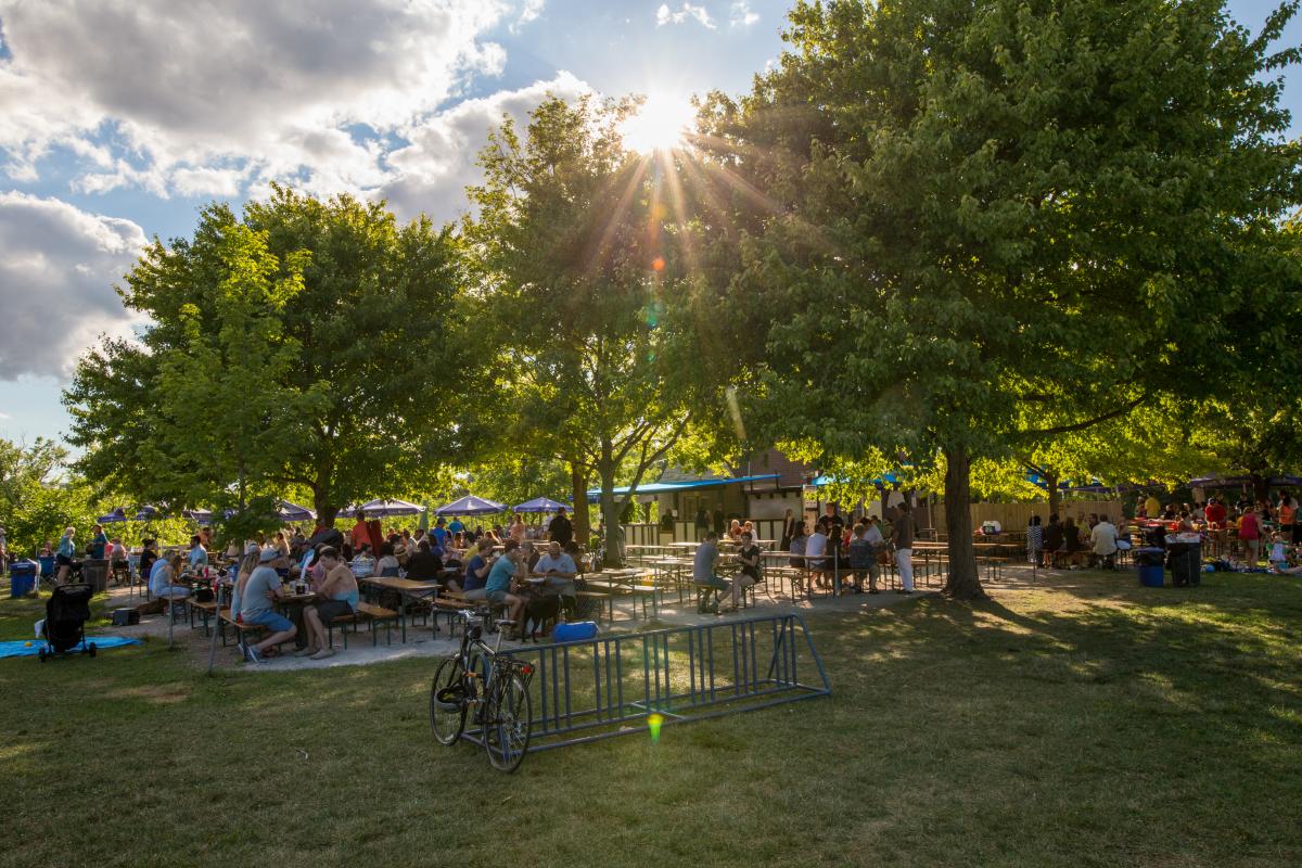 People gathered at picnic tables under shady trees at Estabrook Beer Garden in Milwaukee on a sunny day, with blue umbrellas, strollers, and bikes in the foreground.