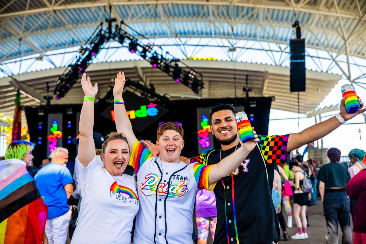 Three people smiling and cheering at Milwaukee PrideFest, wearing rainbow-themed clothing and holding drinks in rainbow koozies, with a colorful "Pride" sign on stage in the background.