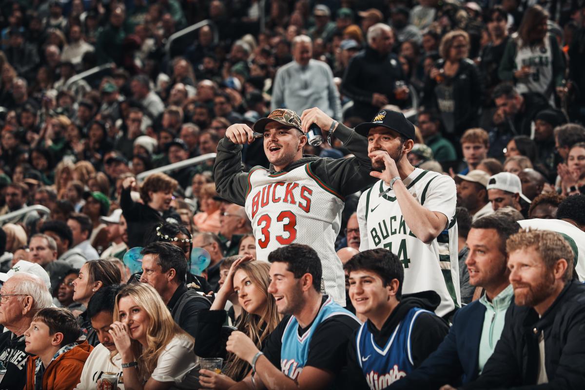 Two excited fans wearing Milwaukee Bucks jerseys cheer from their seats during a packed game at Fiserv Forum, surrounded by a crowd of fellow spectators watching the action.