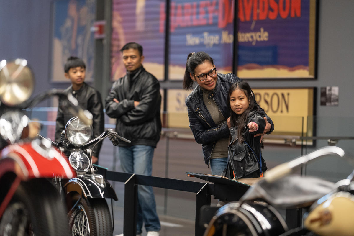 A family of four, all dressed in black leather jackets, visits the Harley-Davidson Museum. A smiling woman and a young girl in the foreground point at a vintage motorcycle display, while two other family members stand behind them observing.