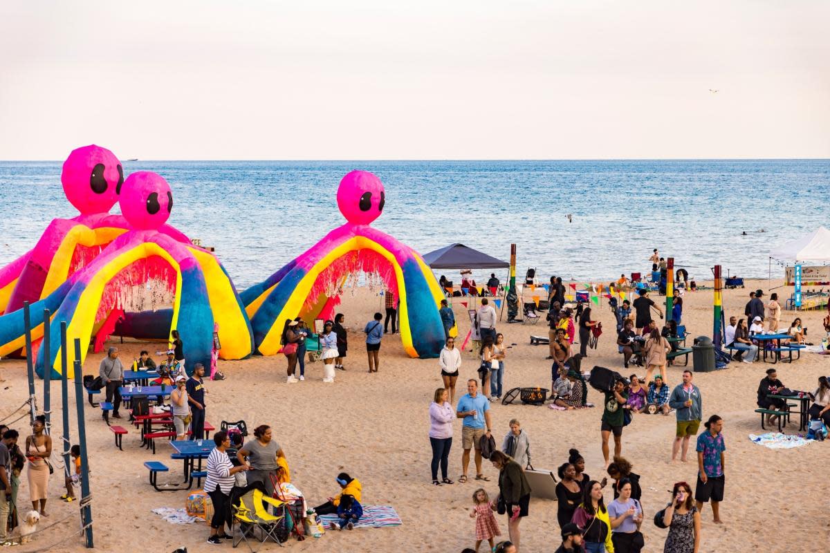 A lively beach festival scene at Milwaukee’s lakefront, featuring three large, colorful inflatable octopus structures with pink heads and rainbow-colored arms. Crowds of people walk, relax, and gather around picnic tables on the sand, with Lake Michigan visible in the background. The event includes vendor tents, painted poles, and beach games, creating a festive, family-friendly atmosphere.