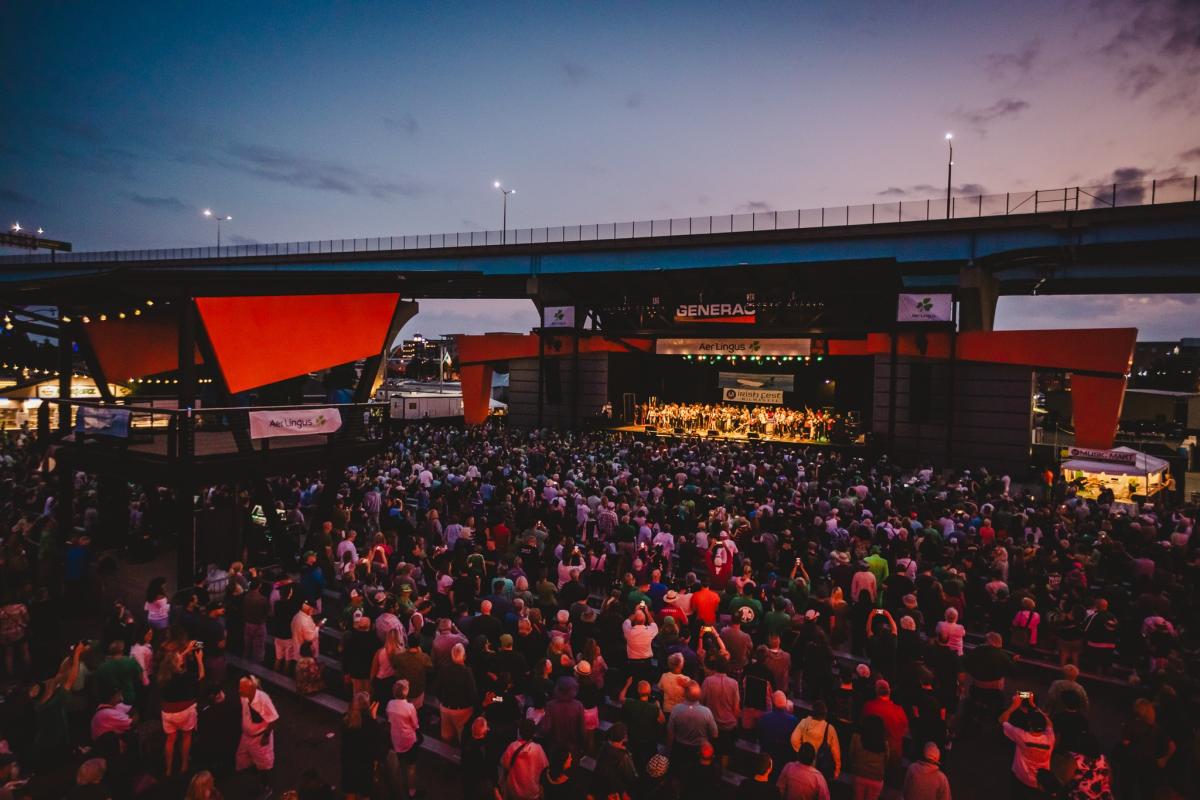 A large crowd gathers at the American Family Insurance Amphitheater during twilight to watch a live performance on stage. The audience fills the venue, with some people standing and others seated. The stage is framed by bold red architectural panels, and overhead signage includes sponsors like Generac and Aer Lingus. The evening sky casts a purple hue as lights from the venue and city create a festive atmosphere. A banner reading “Irish Fest” is visible on the stage backdrop.