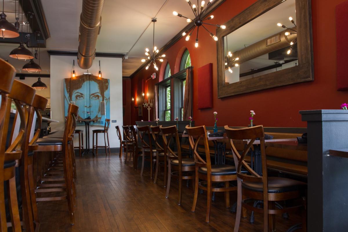A cozy, upscale restaurant interior with warm red walls, wooden floors, and modern chandelier lighting. Wooden chairs line long tables topped with small flower vases, and a large mirror reflects the room’s lighting. A prominent blue-toned portrait of a woman hangs on a white wall at the far end, flanked by two high-top chairs beneath an exposed duct ceiling.
