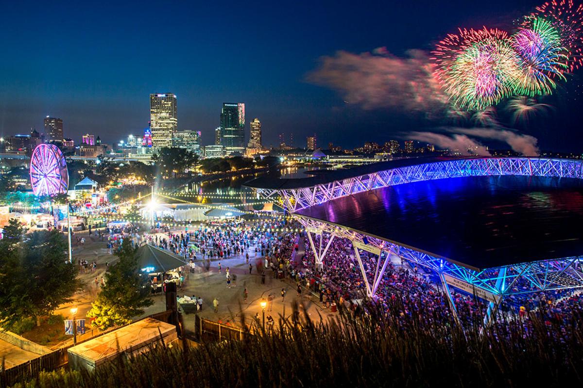 A vibrant nighttime scene at Summerfest grounds in Milwaukee, featuring a large crowd, illuminated Ferris wheel, and the glowing roof of the American Family Insurance Amphitheater. The downtown skyline sparkles in the background as colorful fireworks burst over Lake Michigan, adding to the festive atmosphere.